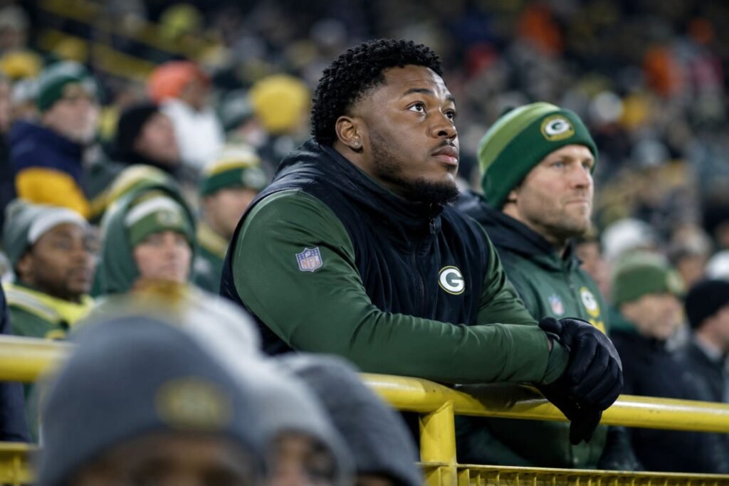 Josh Jacobs watching an NFL game from the Green Bay Packers sideline in a crowded stadium